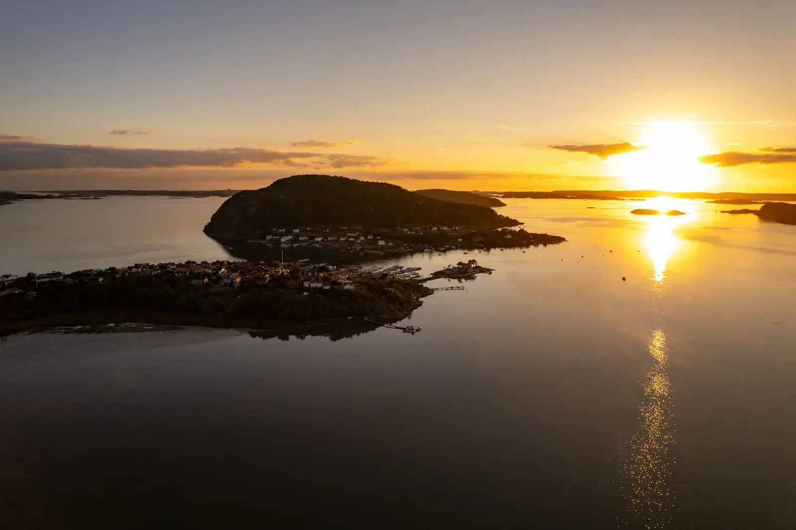 Norwegische Schärenküste von oben im Abendlicht: Eine bewaldete Insel liegt als dunkle Silhouette im Gegenlicht, die Sonne zeichnet eine helle Bahn über das ruhige Wasser.