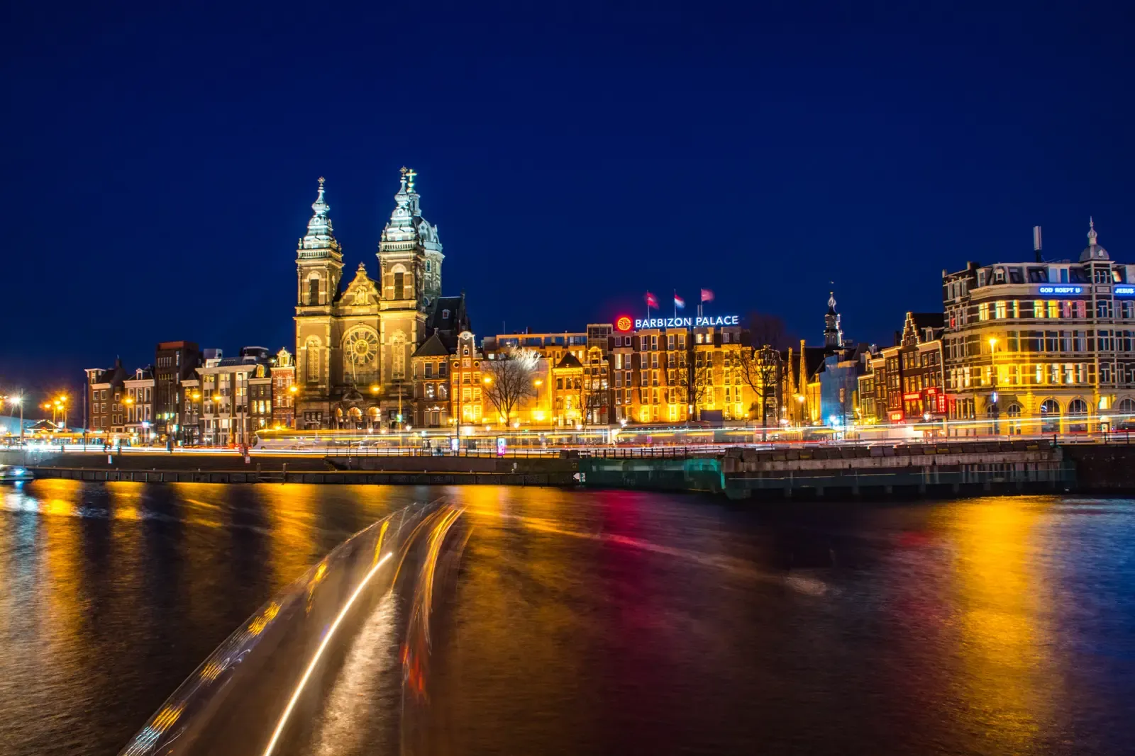 Amsterdam in der blauen Stunde: die beleuchtete Sint-Nicolaaskerk am Wasser, davor zieht ein Boot eine helle Lichtspur über den Kanal.