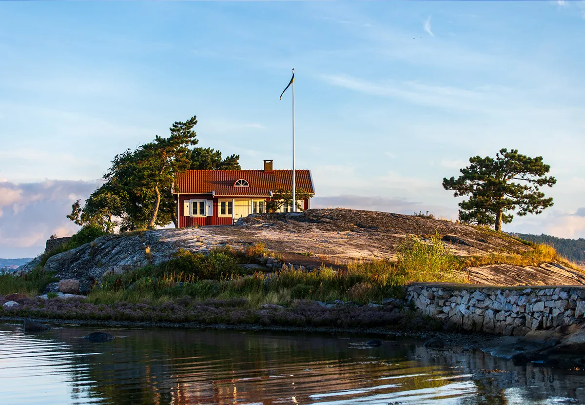 Rotes Holzhaus mit norwegischer Flagge auf einer Felsinsel am Wasser.