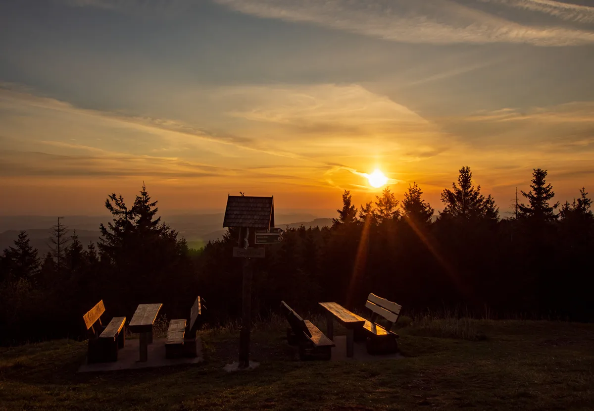 Sonnenaufgang über bewaldeten Hügeln, im Vordergrund Bänke und ein Wegweiser.