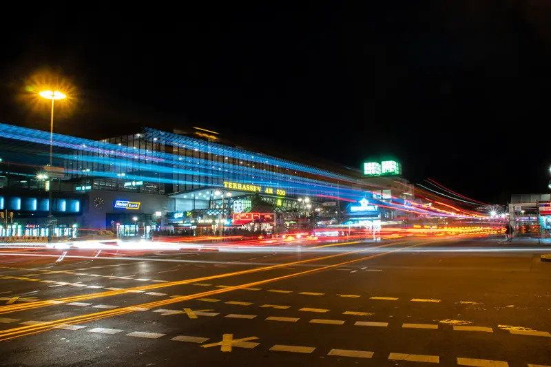 Kreuzung bei Nacht mit langen Lichtspuren, dahinter ein beleuchteter Bahnhof.