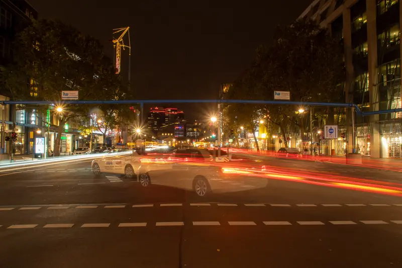 Nächtliche Straße mit Lichtspuren, im Hintergrund Baukräne.