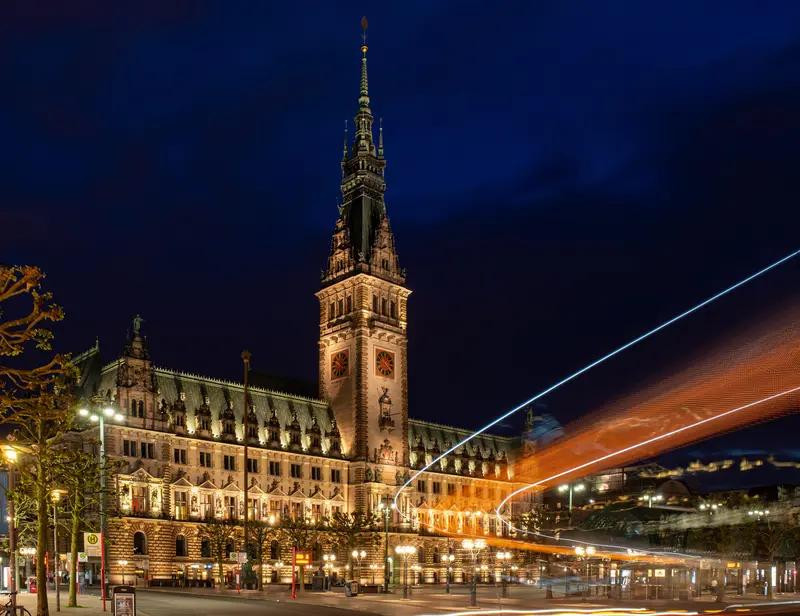 Das Hamburger Rathaus in der blauen Stunde, davor eine helle Lichtspur.
