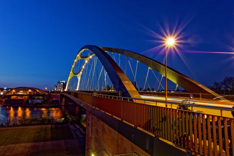 Die Osthafenbrücke in Frankfurt in der blauen Stunde. An der Straßenlaterne rechts steht ein vielstrahliger Blendenstern, kleinere Sterne sitzen an den Lichtern des Brückenbogens.