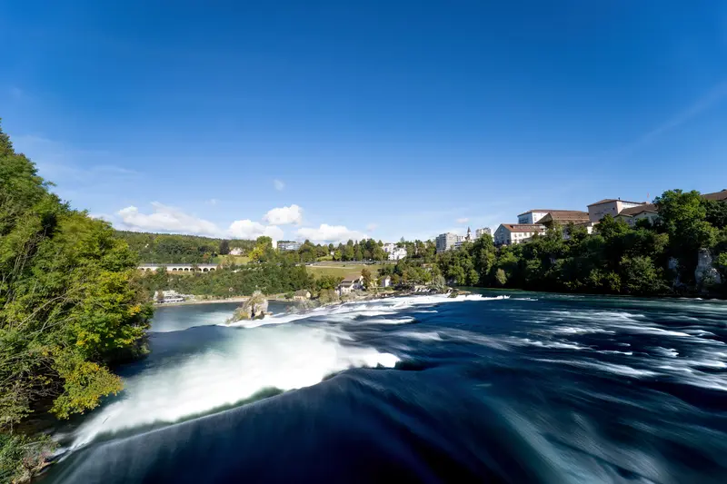 Der Rheinfall von oben gesehen, das Wasser zieht als breite weiße Bahn durch das Bild, dahinter bewaldete Ufer und Ortsbebauung.