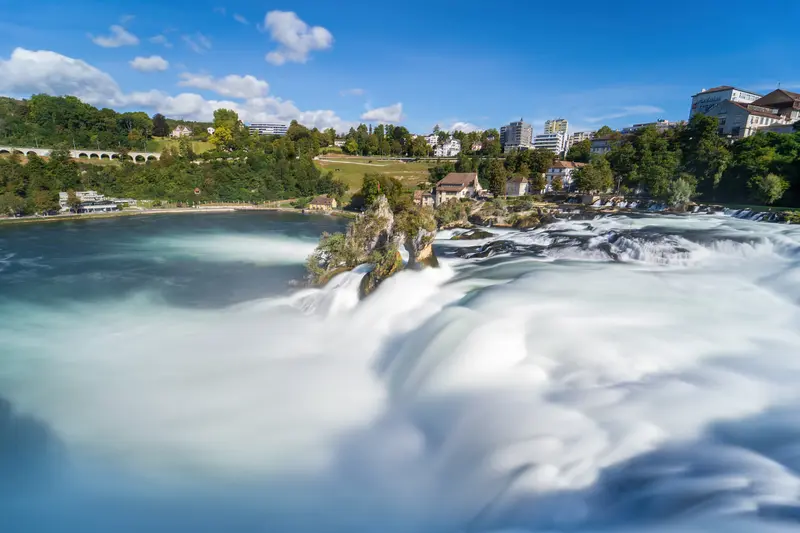 Der Rheinfall mit türkisfarbenem Becken, mitten im Strom der bewachsene Felsen, dahinter die Uferbebauung.