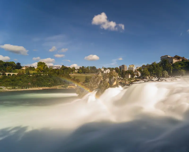Der Rheinfall im Gegenlicht, über der aufsteigenden Gischt steht ein Regenbogen.