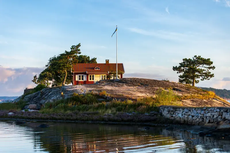 Rotes Holzhaus mit Fahnenmast auf einer Felsinsel, davor stilles Wasser.