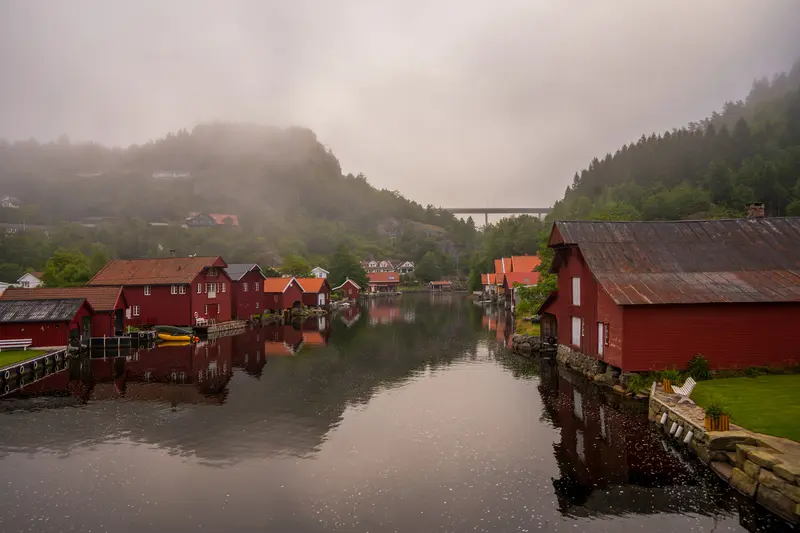 Bootshäuser am Wasser im Morgennebel, gespiegelt in der stillen Oberfläche.