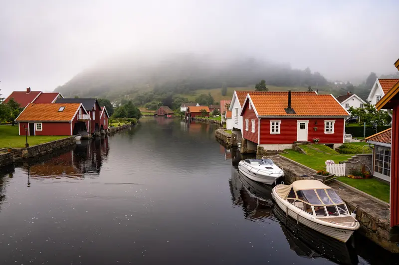 Rote Bootshäuser und Boote an einem Kanal, Nebel über den Hügeln dahinter.