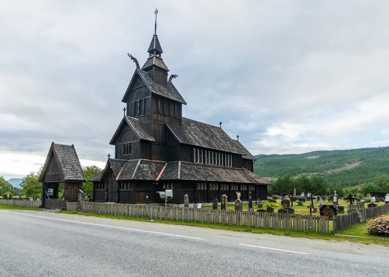 Stabkirche aus dunklem Holz mit Friedhof, dahinter bewaldete Hänge.