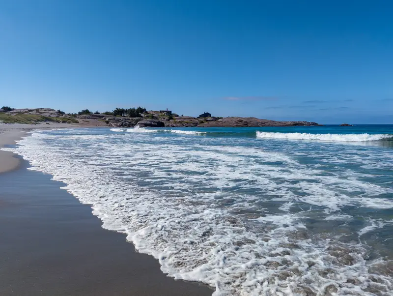 Brandung läuft auf einen Sandstrand, dahinter Felsen und offenes Meer.