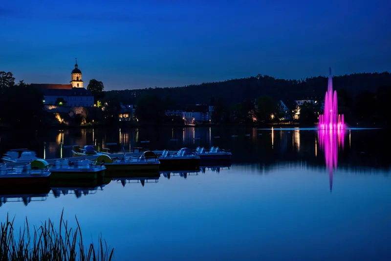 Blaue Stunde am See, eine magentafarbene Fontäne, davor liegen Tretboote.