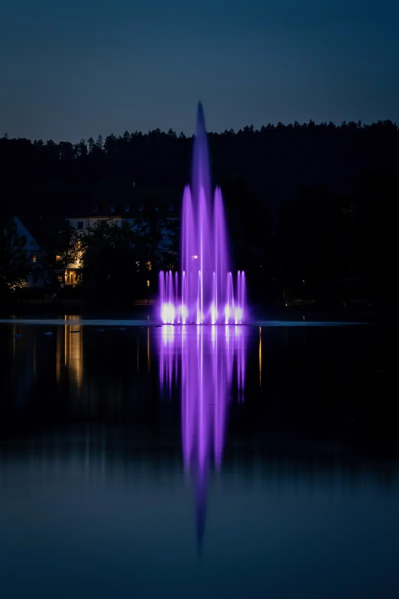 Beleuchtete Wasserfontäne in Violett auf dem dunklen See.