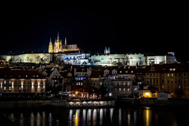 Die Prager Burg auf dem Hügel bei Nacht, davor die Moldau und die Uferbebauung.